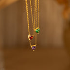 Three Heart Birthstone Necklaces with heart-shaped red, green, and purple stones hang against a blurred brown background—an ideal personalized jewelry gift for birthdays.
