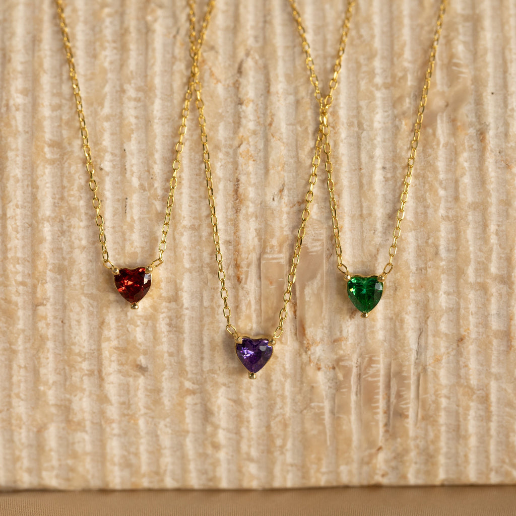 Three Heart Birthstone Necklaces with red, purple, and green stones rest on a textured beige surface—a stunning personalized gift perfect for birthdays.