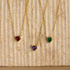 Three Heart Birthstone Necklaces with red, purple, and green stones rest on a textured beige surface—a stunning personalized gift perfect for birthdays.