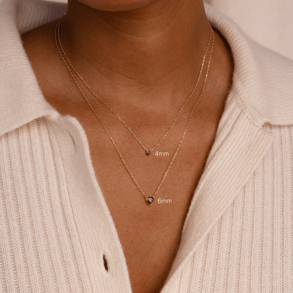 Two gold chain necklaces with small pendants, featuring a 4mm and 6mm Heart Birthstone Necklace, layered over a cream ribbed sweater—personalized jewelry that's perfect for a thoughtful birthday gift.