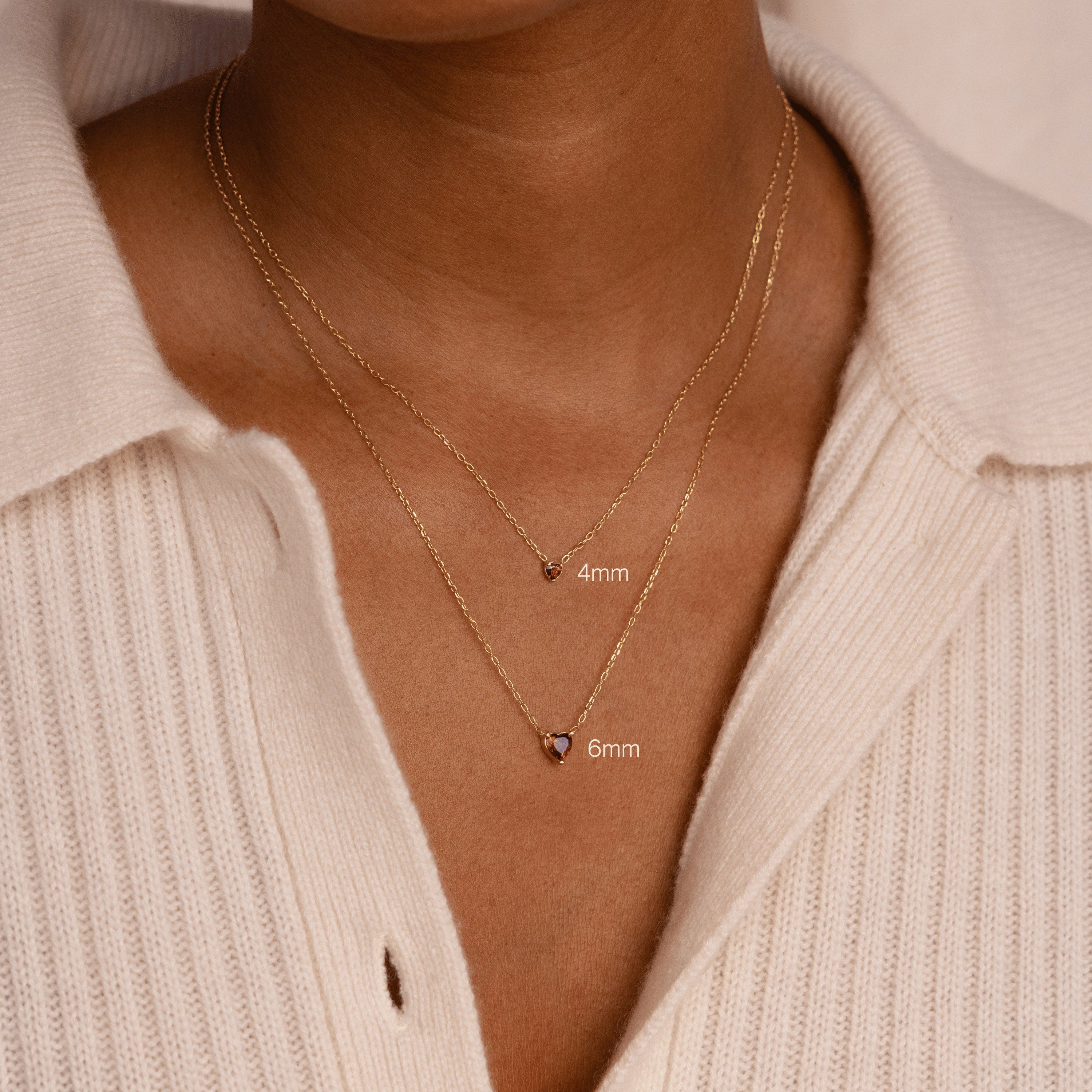 Two gold chain necklaces with small pendants, featuring a 4mm and 6mm Heart Birthstone Necklace, layered over a cream ribbed sweater—personalized jewelry that's perfect for a thoughtful birthday gift.