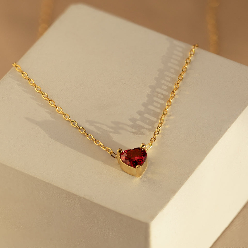 The Heart Birthstone Necklace features a vibrant red gemstone pendant, elegantly showcased on a light-colored geometric block—a perfect birthday gift or timeless personalized jewelry piece.