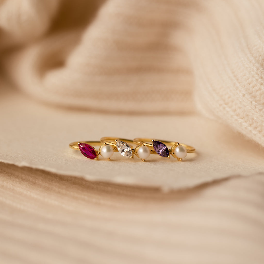 The Marquise Birthstone & Pearl Ring, featuring gold with colorful gemstones and pearls, is displayed on a soft beige textured fabric background.