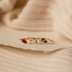 The Marquise Birthstone & Pearl Ring, featuring gold with colorful gemstones and pearls, is displayed on a soft beige textured fabric background.