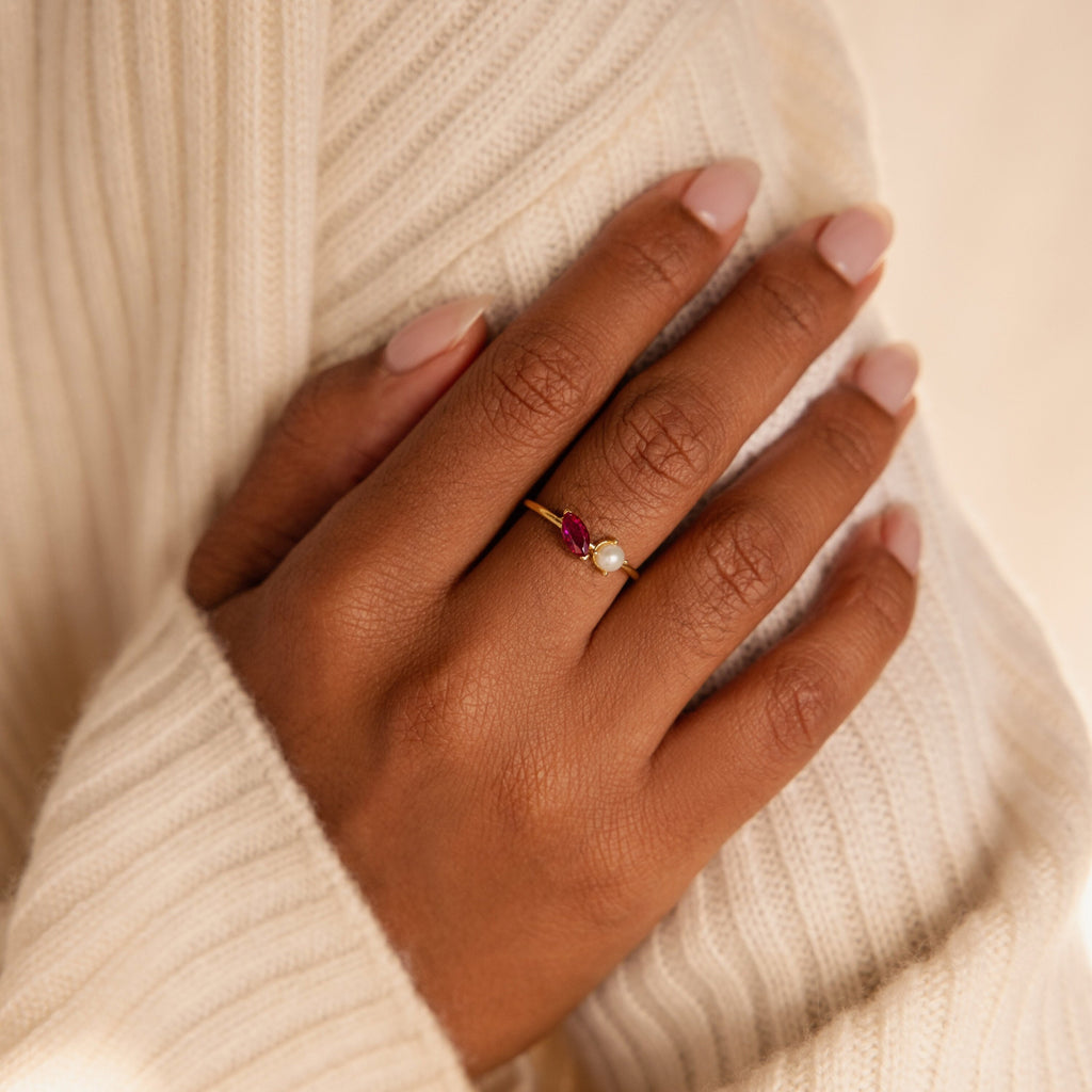 A hand with nude nails wears the Marquise Birthstone & Pearl Ring, featuring a red gemstone and pearl, resting on a cream sweater.