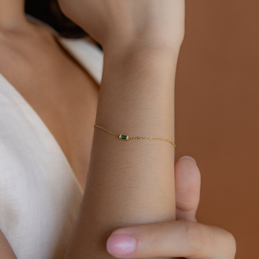 The Baguette Birthstone Bracelet with a gold chain and a small green stone shines on a woman's wrist against a neutral background.