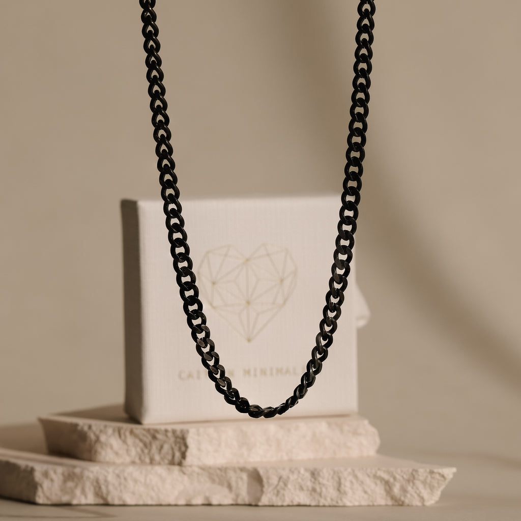 The Men's Black Curb Chain is displayed in front of a white box with a geometric heart design on stone slabs—perfect for men who want to make a bold statement.