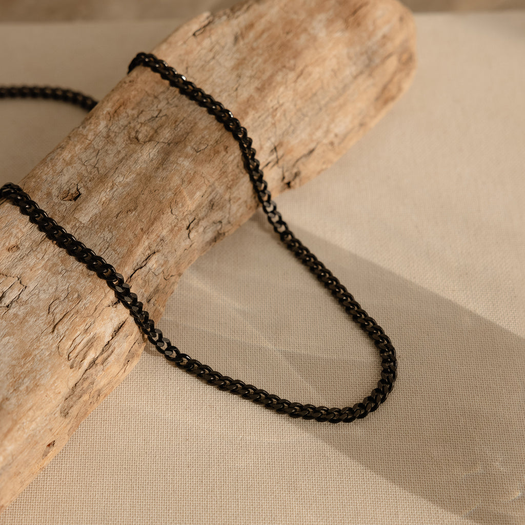 The Men's Black Curb Chain is displayed on light textured wood atop beige fabric, creating a bold statement necklace and a perfect gift for men.