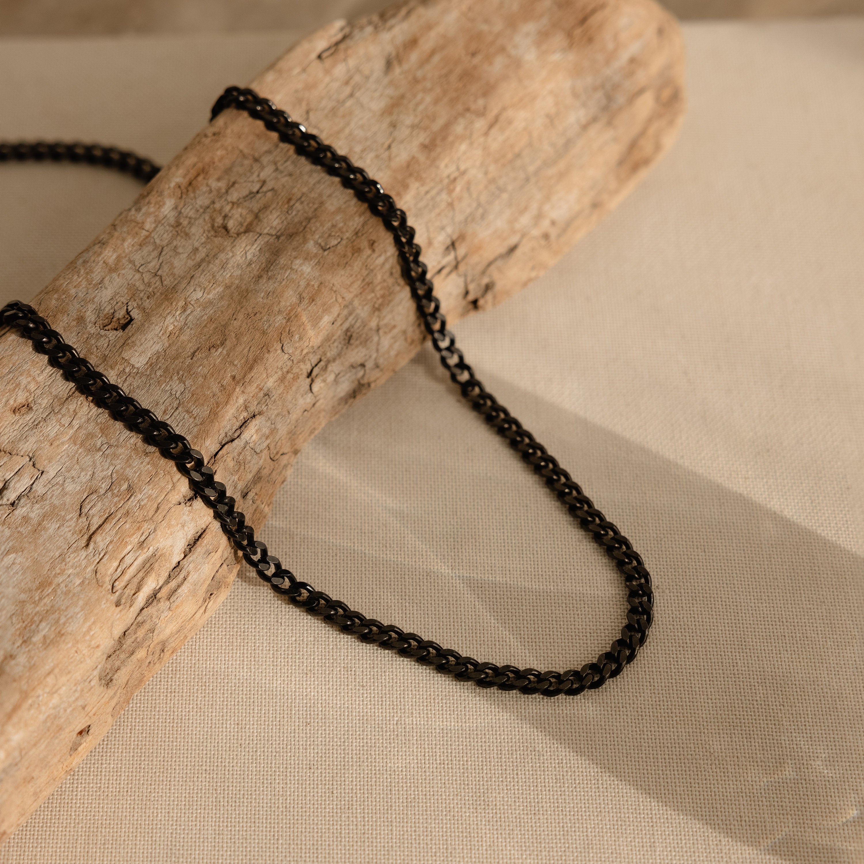 The Men's Black Curb Chain is displayed on light textured wood atop beige fabric, creating a bold statement necklace and a perfect gift for men.