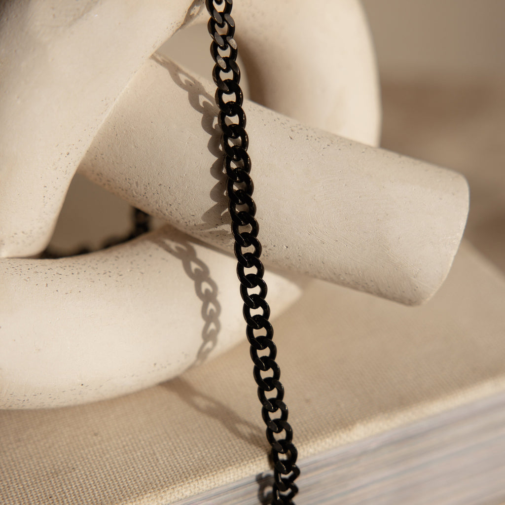 The Men's Black Curb Chain, displayed on an abstract white ceramic sculpture atop a textured surface, is a striking statement necklace and a distinctive gift choice for men.