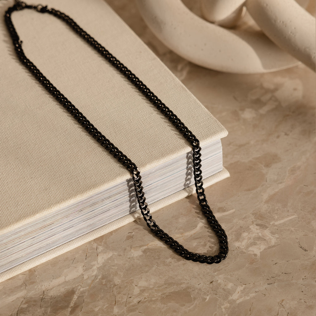 The Men's Black Curb Chain rests on a closed beige book atop a marble surface, making it an ideal gift for men or a bold statement necklace.