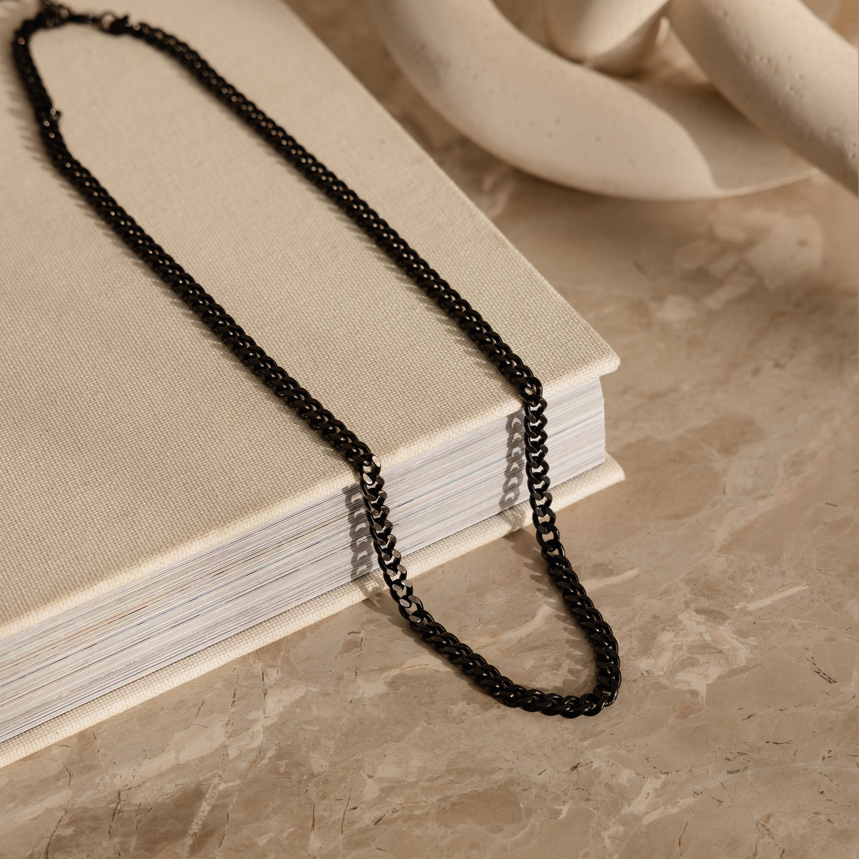 The Men's Black Curb Chain rests on a closed beige book atop a marble surface, making it an ideal gift for men or a bold statement necklace.