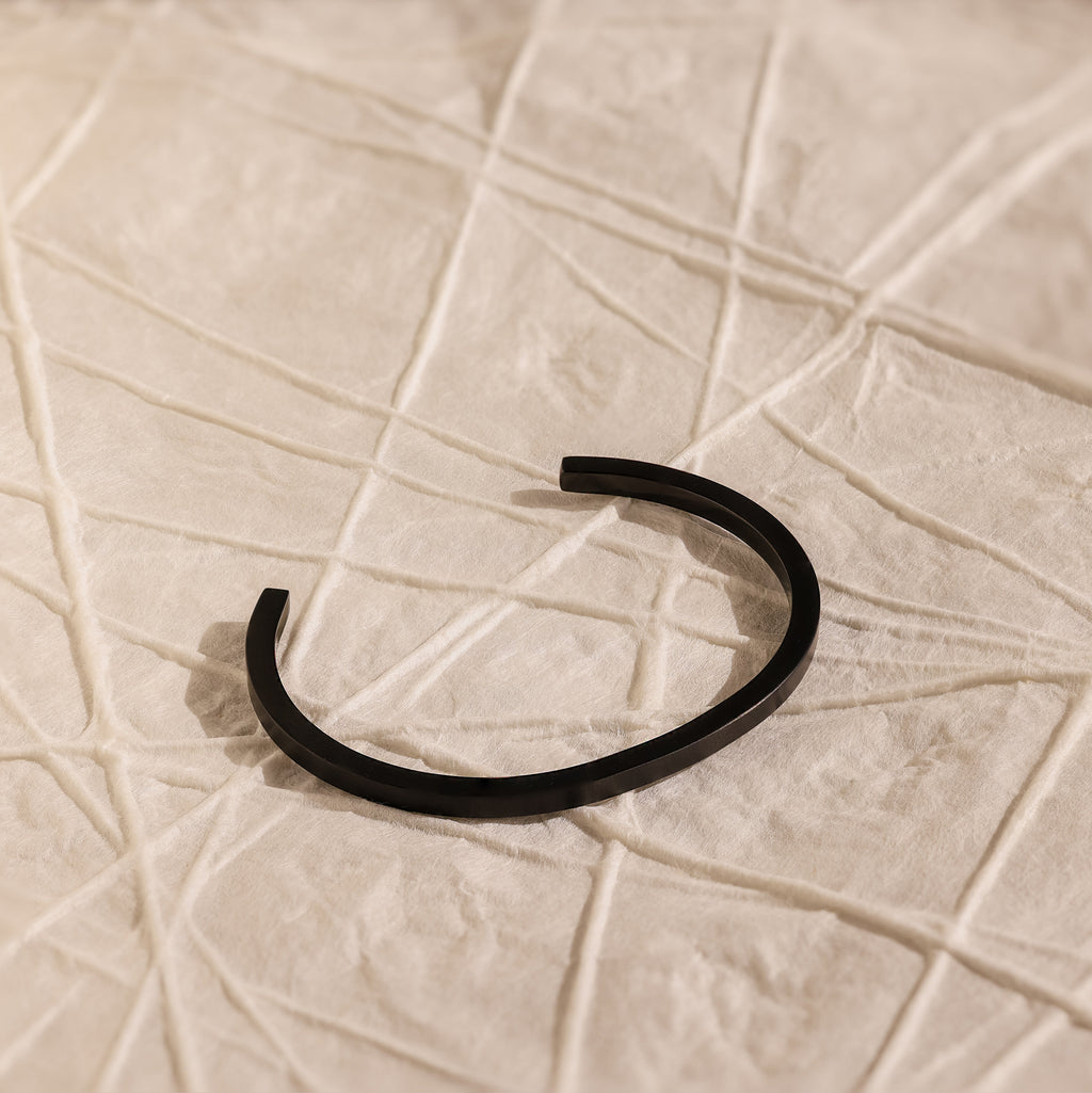 The Men's Black Cuff Bracelet sits on textured, light-toned fabric with geometric lines, offering a modern and stylish gift idea for him.