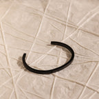 The Men's Black Cuff Bracelet sits on textured, light-toned fabric with geometric lines, offering a modern and stylish gift idea for him.