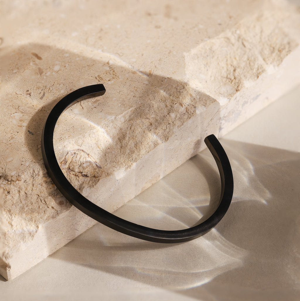 The Men's Black Cuff Bracelet lies on a textured beige stone surface with gentle natural shadows—making it a sleek, thoughtful gift for him.