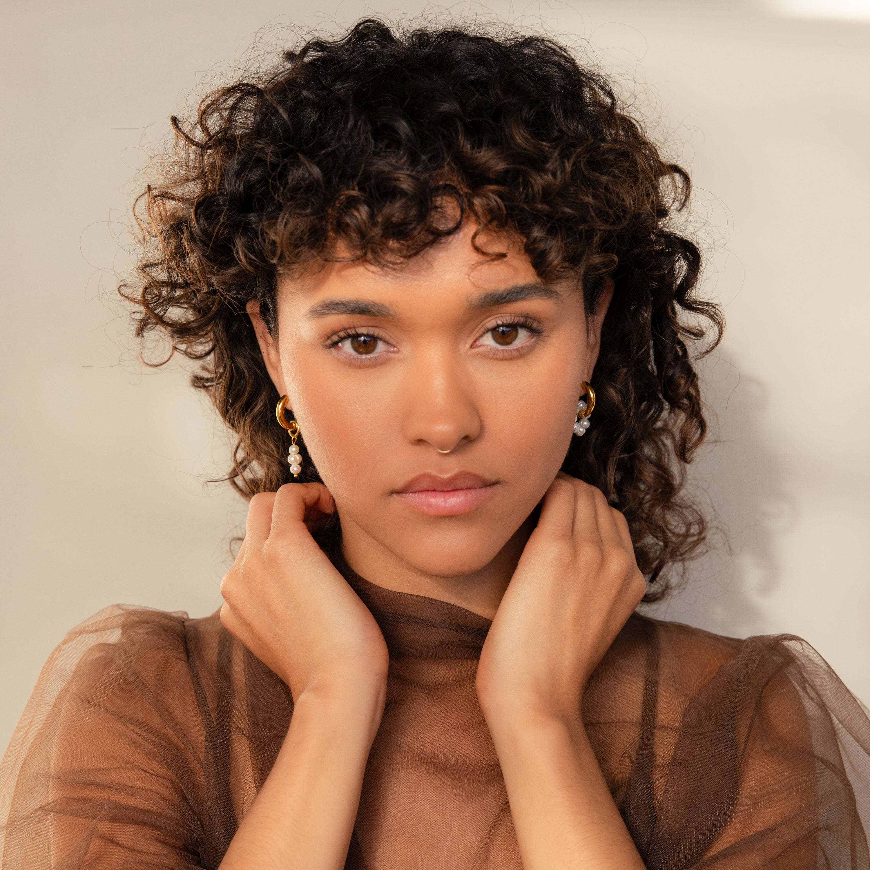 A woman with curly hair and gold earrings poses with her hands near her neck, wearing a sheer brown top and the Mismatched Pearl Drop Hoops.