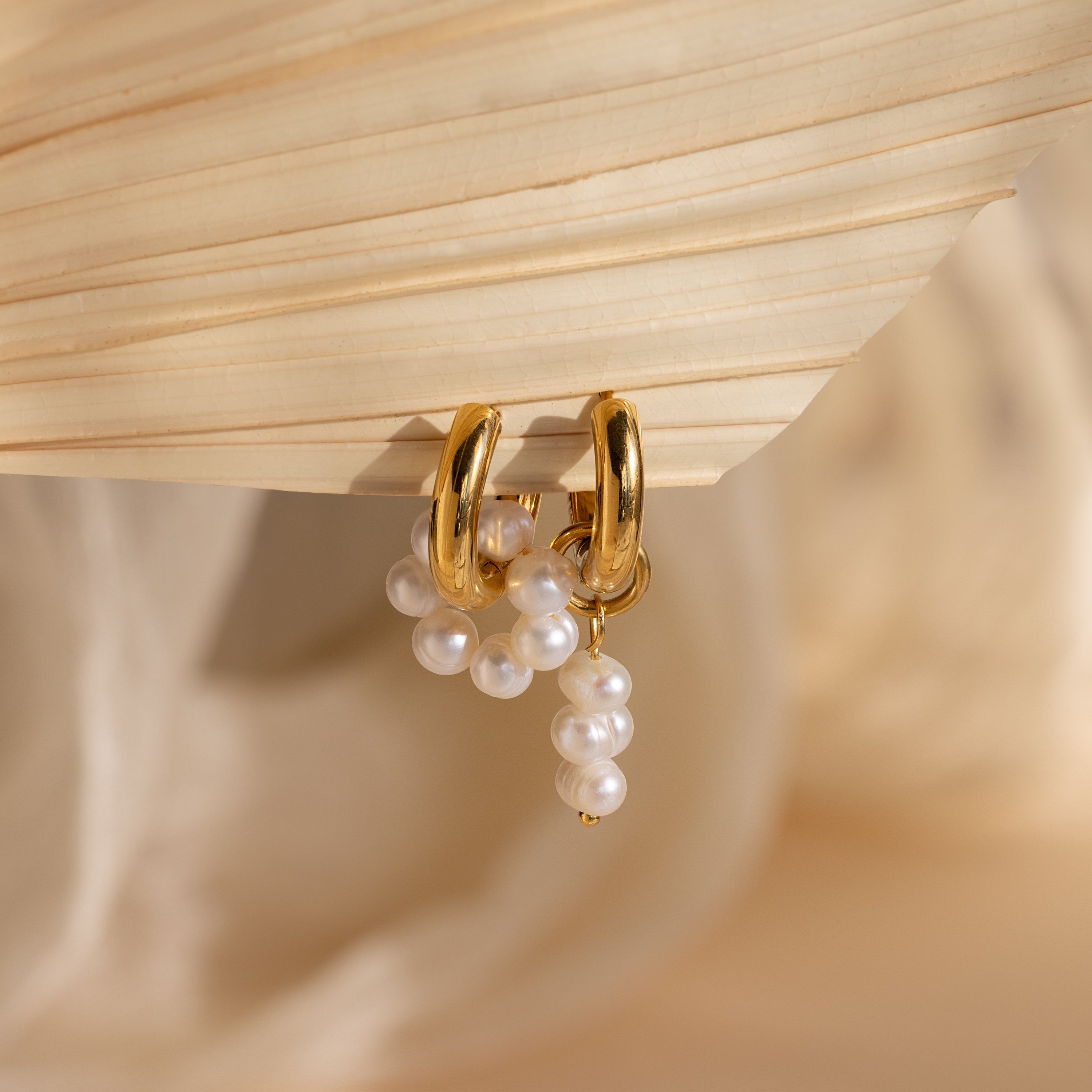 Mismatched Pearl Drop Hoops feature gold hoops with dangling clusters of white pearls, creating a striking statement look, elegantly displayed on a textured beige surface.