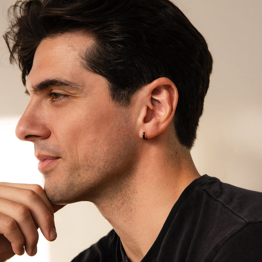 A man with dark hair and an earring, wearing a black shirt, gazes thoughtfully left—highlighting the Men's Black Hoops Set, a stylish pair of earrings perfect as a gift for him.