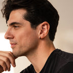 A man with dark hair and an earring, wearing a black shirt, gazes thoughtfully left—highlighting the Men's Black Hoops Set, a stylish pair of earrings perfect as a gift for him.