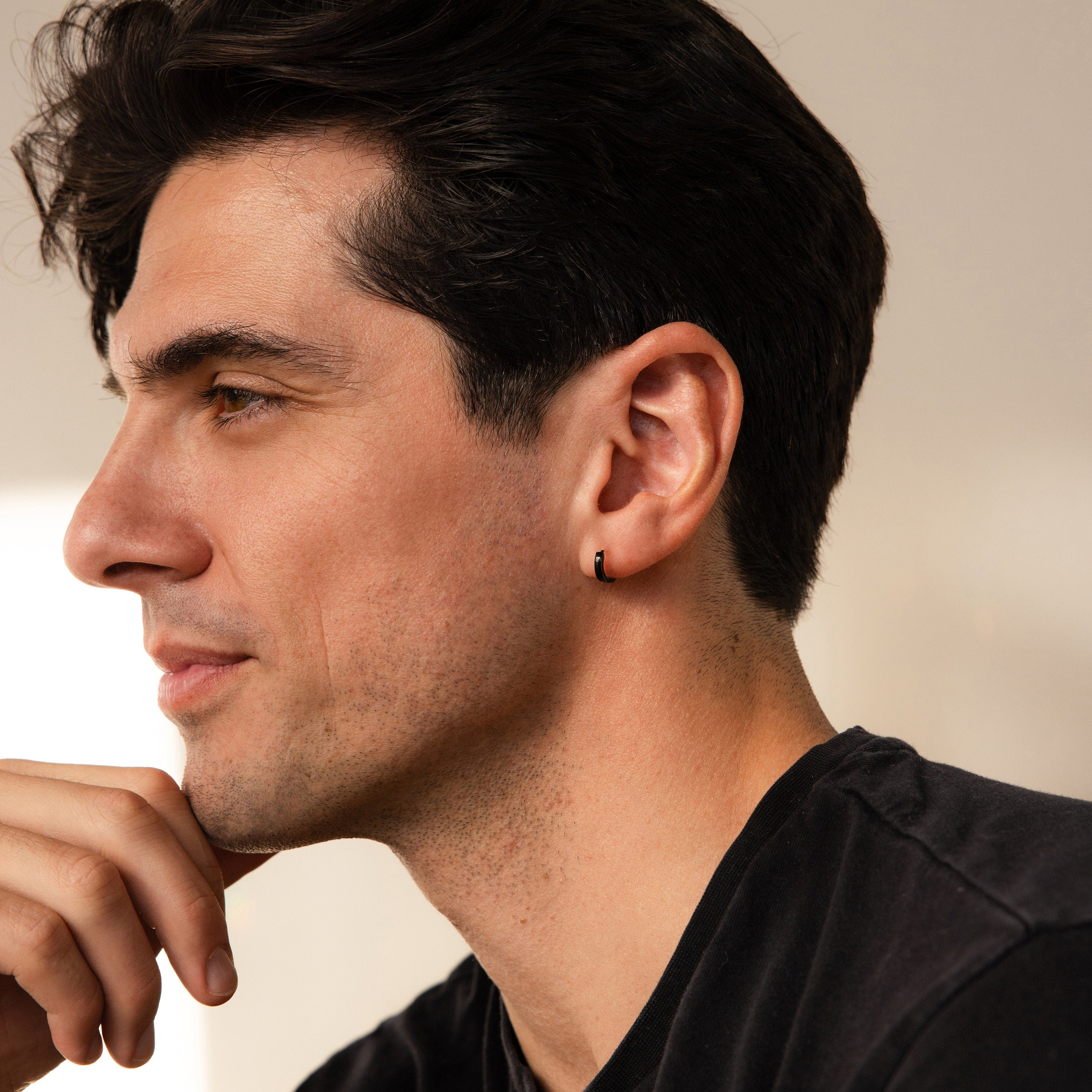 A man with dark hair and an earring, wearing a black shirt, gazes thoughtfully left—highlighting the Men's Black Hoops Set, a stylish pair of earrings perfect as a gift for him.
