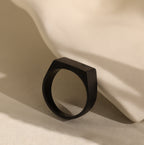 Men's Black Signet Ring with a matte geometric design, placed against a smooth, light surface with soft shadows.