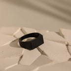 The Men's Black Signet Ring is displayed on a textured, off-white surface with soft lighting and gentle shadows.