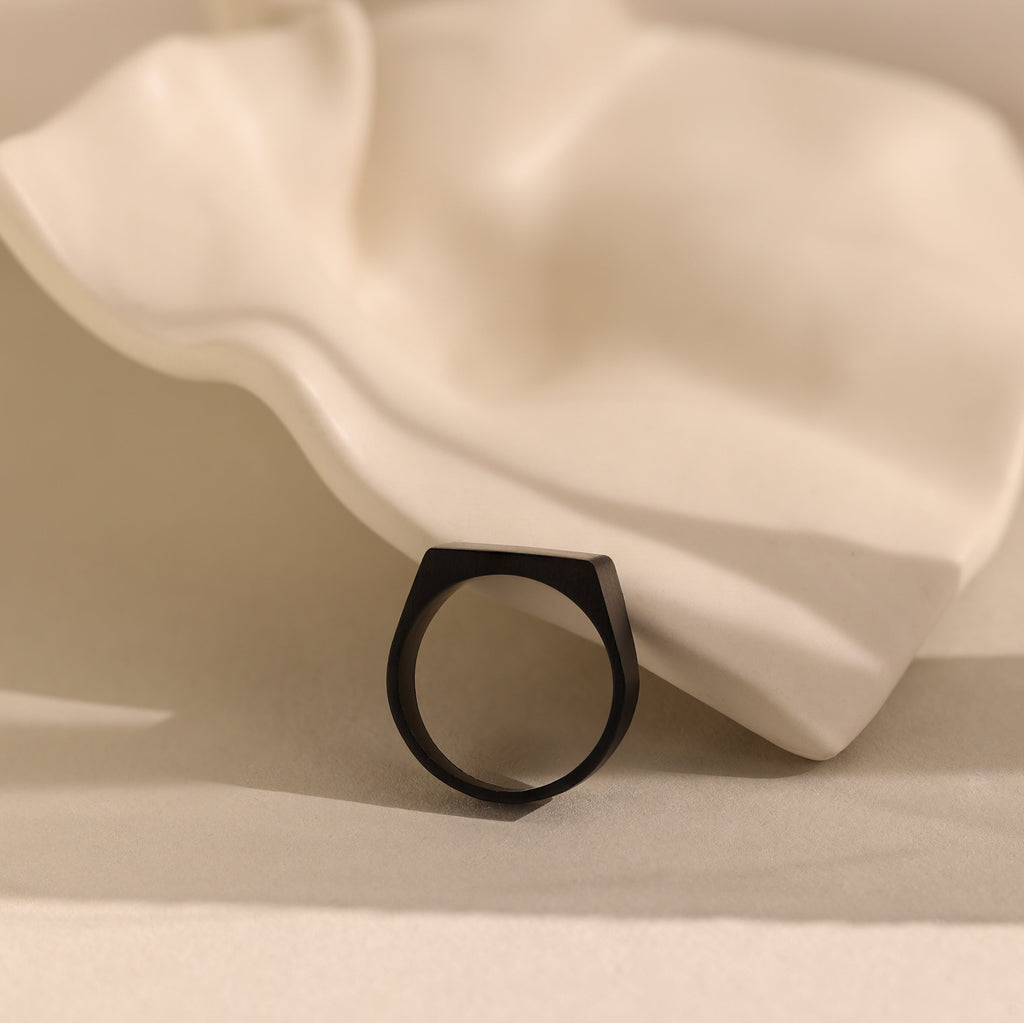 The Men's Black Signet Ring rests on a wavy white ceramic surface with a neutral background.