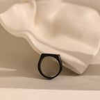 The Men's Black Signet Ring rests on a wavy white ceramic surface with a neutral background.