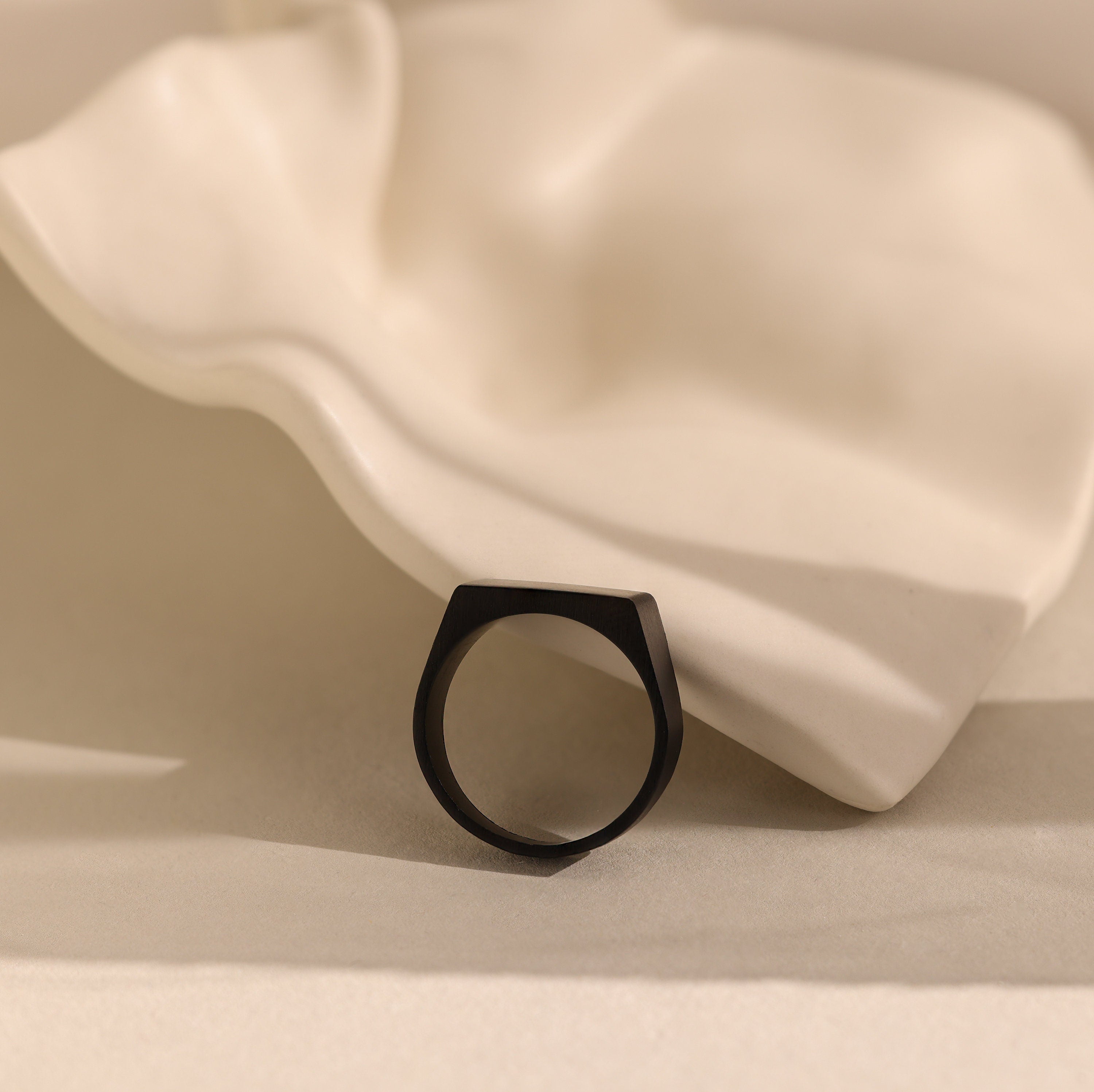 The Men's Black Signet Ring rests on a wavy white ceramic surface with a neutral background.