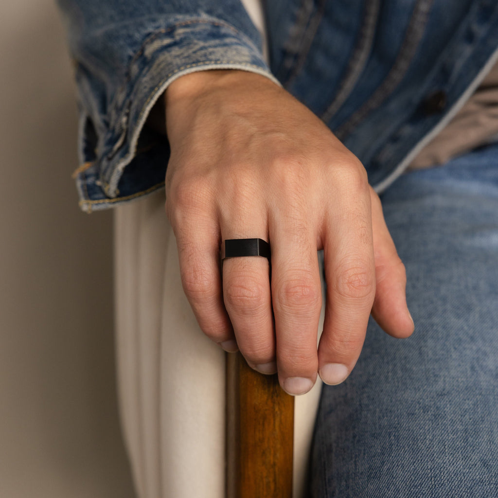 A man wearing the Men's Black Signet Ring rests his hand on a wooden armrest, dressed in denim.