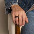 A man wearing the Men's Black Signet Ring rests his hand on a wooden armrest, dressed in denim.