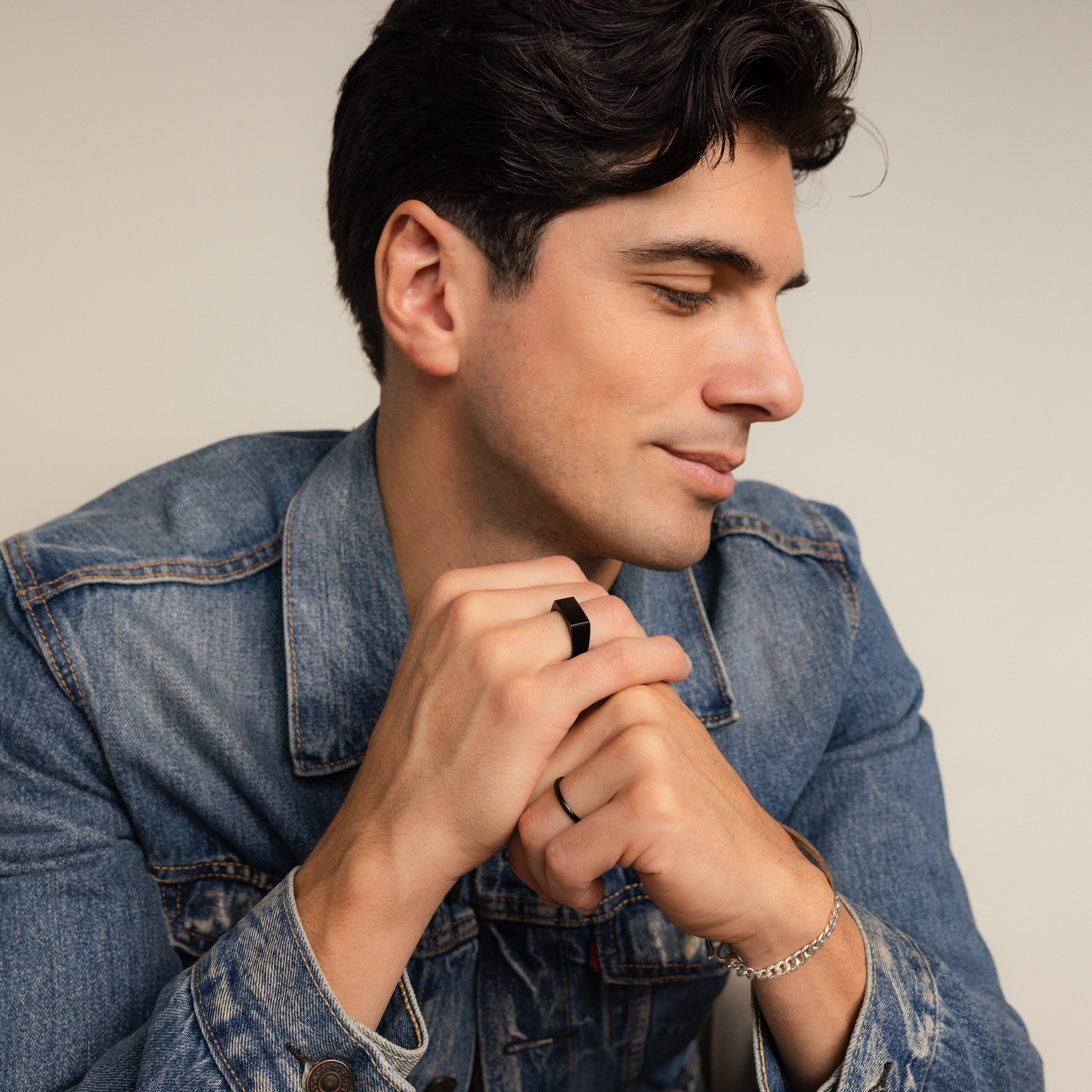 A man with dark hair in a denim jacket rests his chin on his clasped hands, wearing the Men's Black Signet Ring.