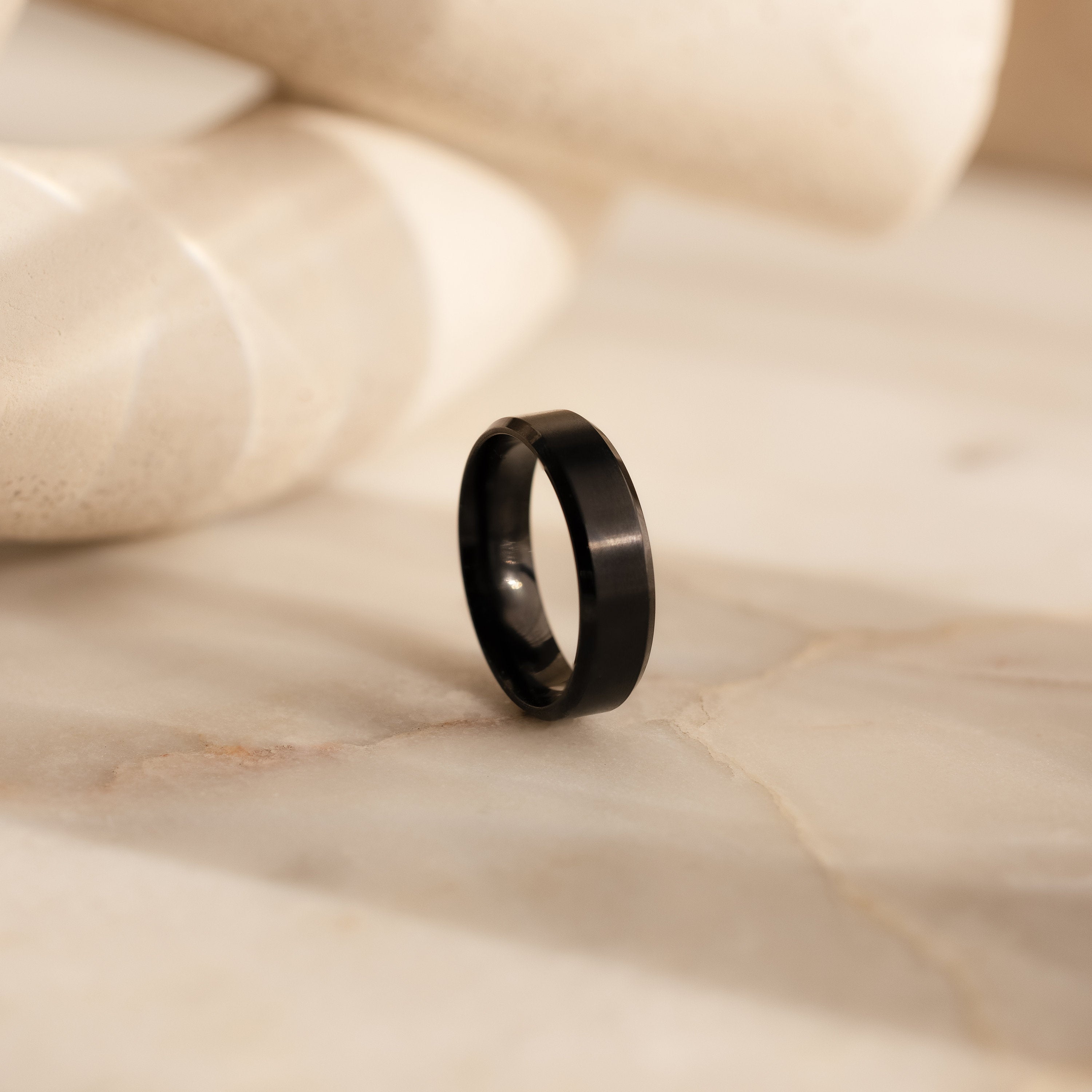 The Men's Thick Black Ring stands upright on a light marble surface with a blurred cream background, highlighting the timeless elegance of this standout men's band.