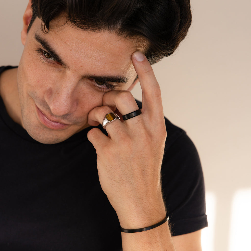A man in a black shirt, looking thoughtful with his hand on his forehead, wears men's rings including the Men's Thick Black Ring and a stylish bracelet.