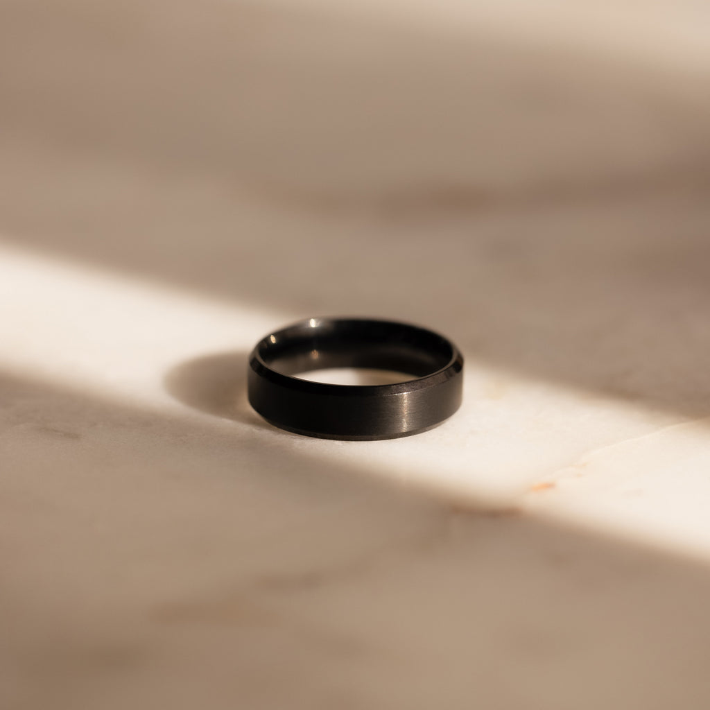 The Men's Thick Black Ring sits on a sunlit, light marble surface with soft shadows—an elegant option for men or as a modern wedding band.