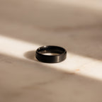 The Men's Thick Black Ring sits on a sunlit, light marble surface with soft shadows—an elegant option for men or as a modern wedding band.
