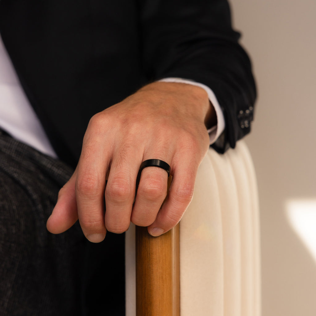A person in a suit sits on a chair, showcasing the Men's Thick Black Ring—a bold black wedding band—on their left hand.