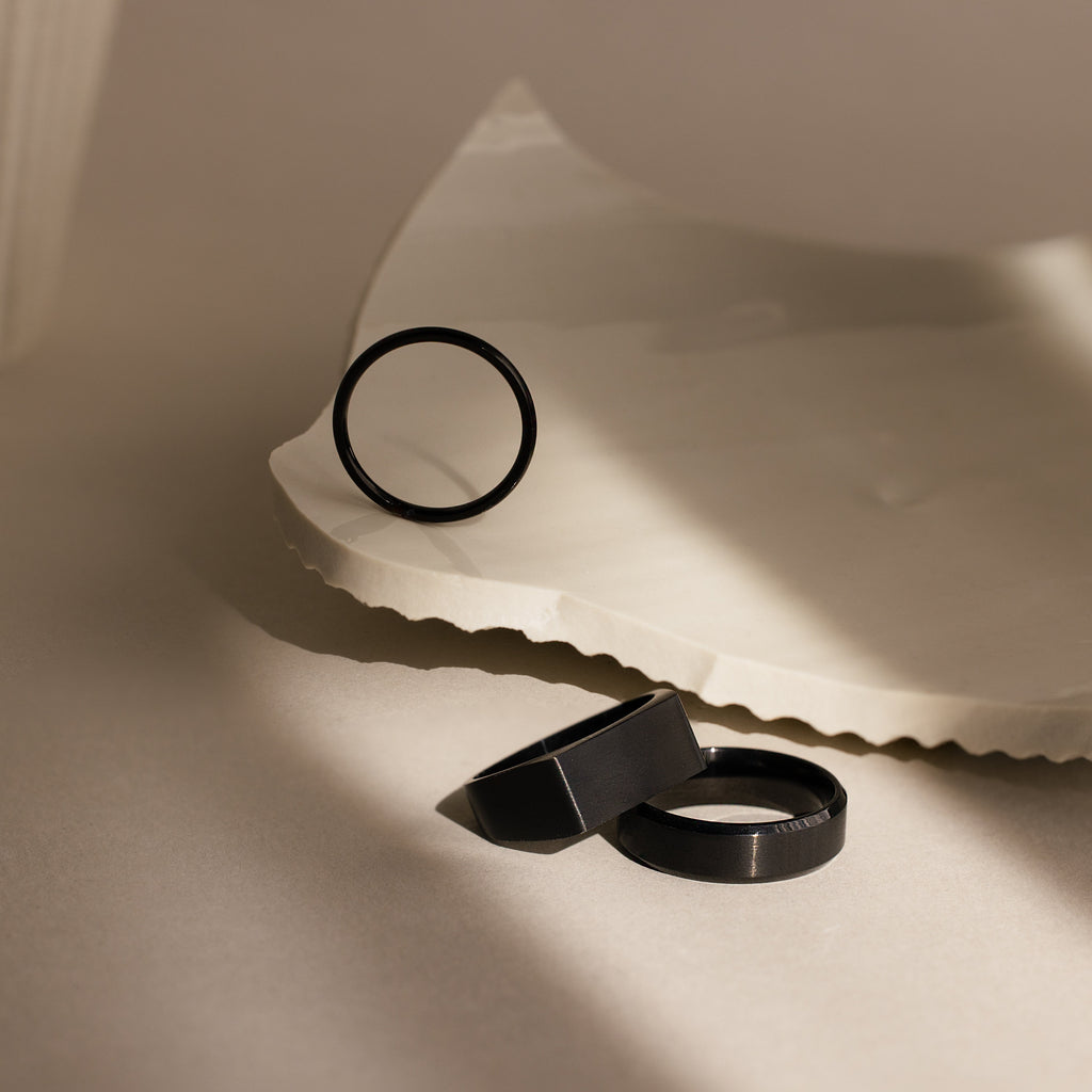 The Men's Black Signet Ring is shown alongside two other black rings on a textured, light-colored surface, highlighted by soft natural lighting and gentle shadows.