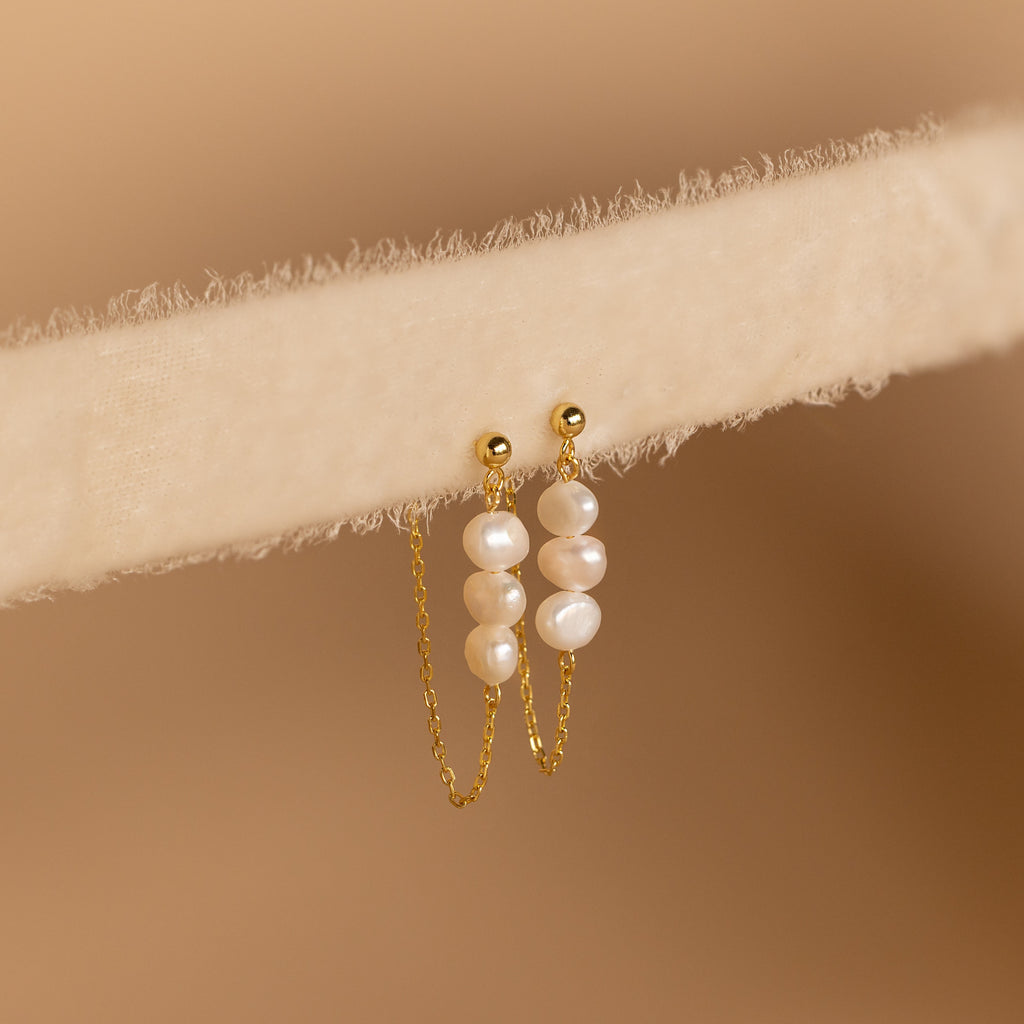 The Triple Pearl Drop Earrings feature delicate chains with small white pearls and rest gracefully on a soft, light beige textured surface—ideal for pearl jewelry lovers.