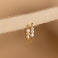 The Triple Pearl Drop Earrings feature delicate chains with small white pearls and rest gracefully on a soft, light beige textured surface—ideal for pearl jewelry lovers.