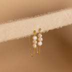The Triple Pearl Drop Earrings feature delicate chains with small white pearls and rest gracefully on a soft, light beige textured surface—ideal for pearl jewelry lovers.