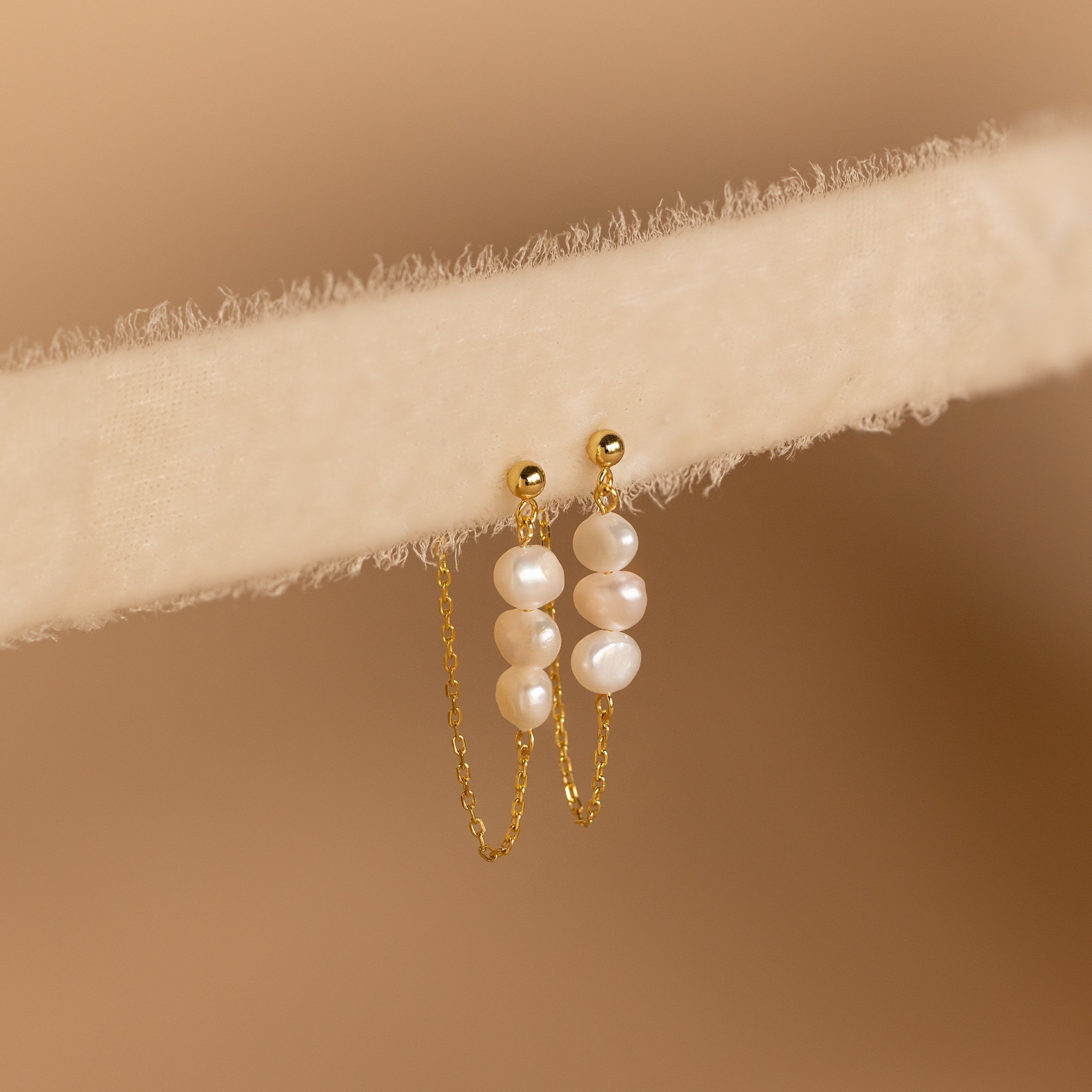 The Triple Pearl Drop Earrings feature delicate chains with small white pearls and rest gracefully on a soft, light beige textured surface—ideal for pearl jewelry lovers.