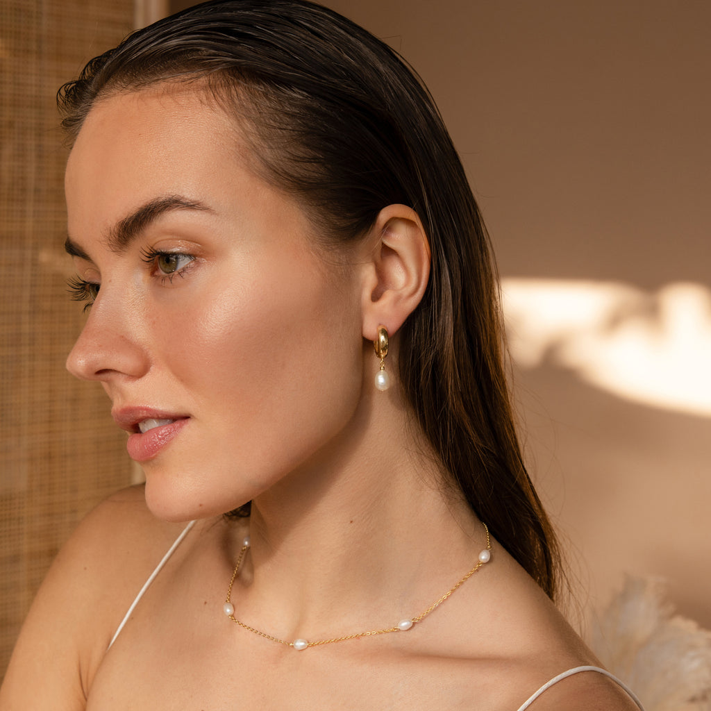A woman with wet, slicked-back hair wears Pearl Drop Hoops and a pearl necklace, bathed in soft natural light.