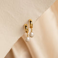 Pearl Drop Hoops feature chunky gold hoops in a minimalist style, accented with dangling white pearls, displayed on a textured beige surface.