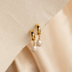 Pearl Drop Hoops feature chunky gold hoops in a minimalist style, accented with dangling white pearls, displayed on a textured beige surface.