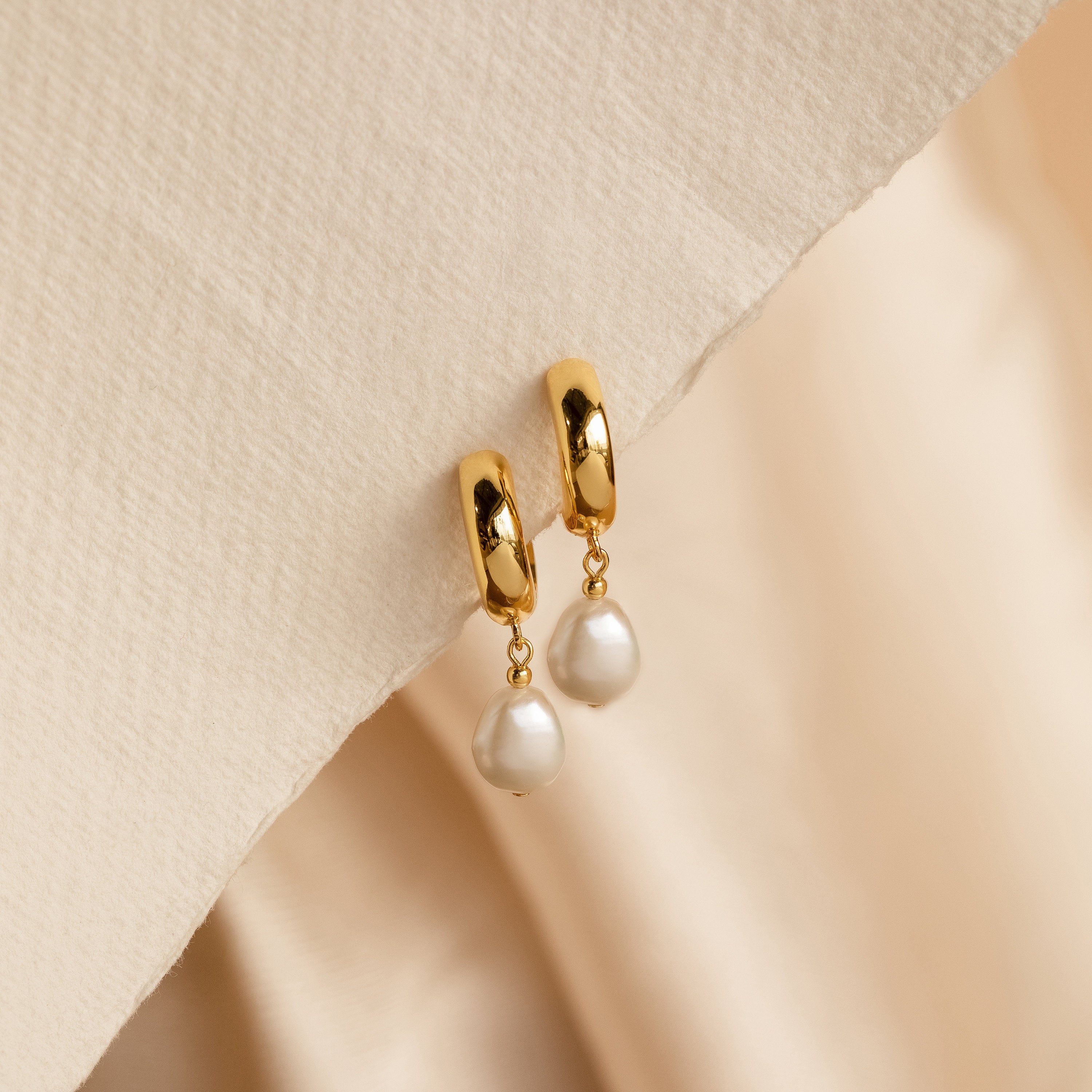 Pearl Drop Hoops feature chunky gold hoops in a minimalist style, accented with dangling white pearls, displayed on a textured beige surface.