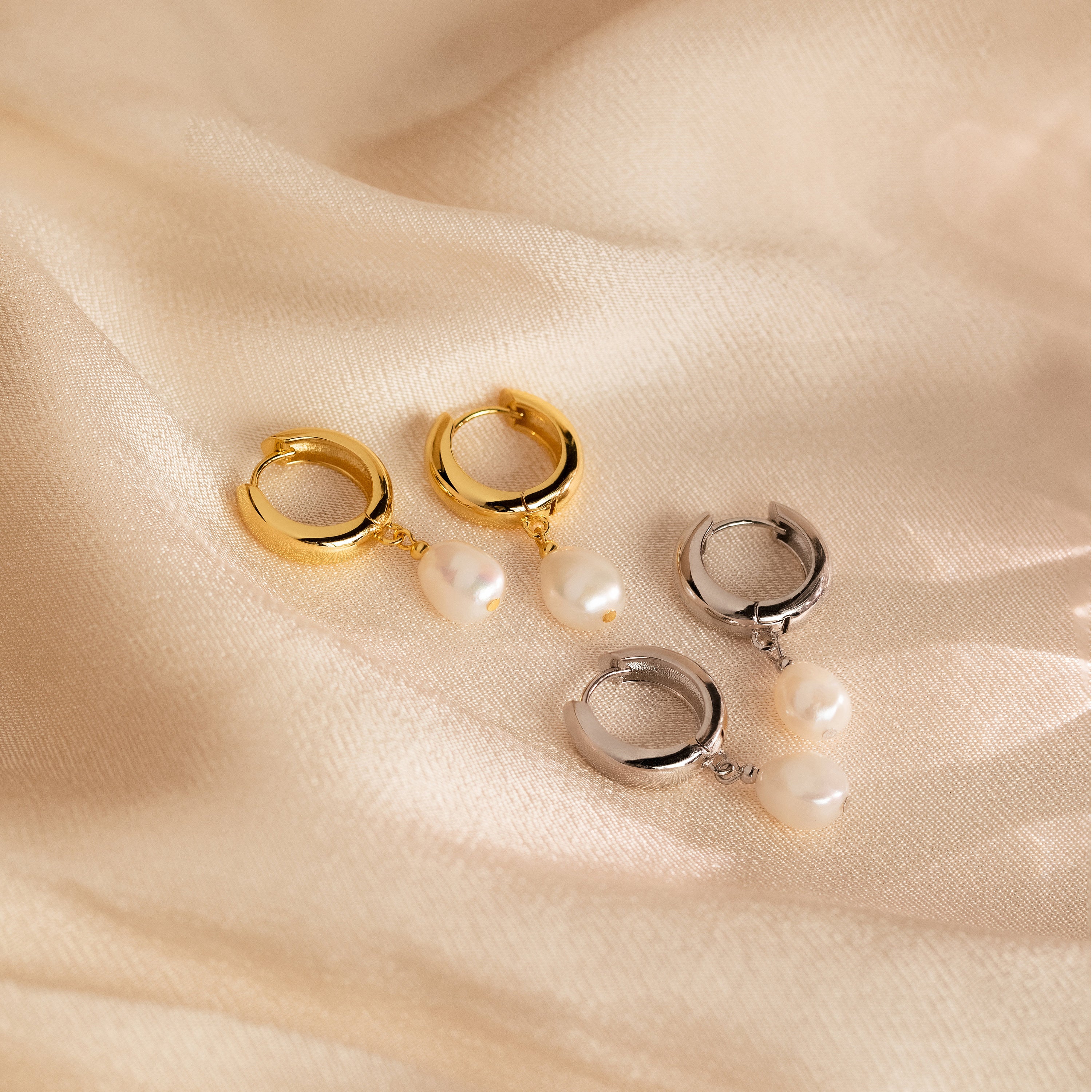 The Pearl Drop Hoops feature a minimalist gold and silver hoop design with delicate pearl drops, beautifully displayed on a soft beige fabric background.