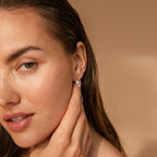 A woman with wet hair touches her Dainty Knot Pearl Earrings, radiating classic charm against a soft beige backdrop.