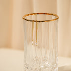 The Pearl Threader Earrings, featuring gold and pearl drops in a minimalistic design, are displayed hanging on the rim of a clear faceted glass against a beige background.