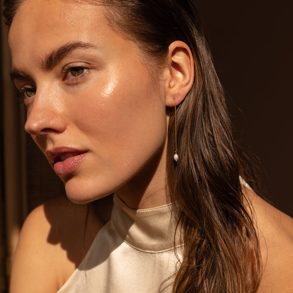 A woman with dewy skin and wet hair glows in soft sunlight, wearing minimalistic Pearl Threader Earrings and a cream halter top.